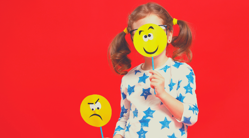 menina de aproximadamente cinco anos segurando uma carinha de cartolina com um smile sorrindo desenhado, e apoiado contra o seu rosto. E na mão direita está segurando um smile de cartolina com um rosto de desapontado desenhado. A foto foi tirada em um fundo vermelho, e a menina está vestida com uma camisa branca com estrelas azuis.