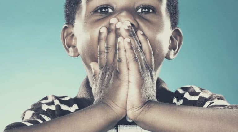Criança negra de aproximadamente quatro anos, com as duas mãos sobre a boca, sinalizando silêncio.