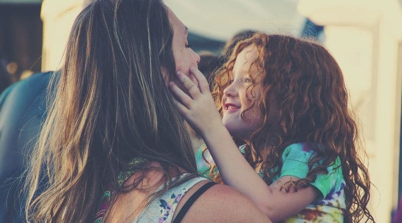 Mãe de aproximadamente trinta e cinco anos, cabelos lisos e castanhos, vestida com uma regata azul segurando sua filha de aproximadamente cinco anos no colo, sua filha é ruiva de cabelos cacheados. segurando o rosto da mãe com ternura e segurança, sorrindo para ela.