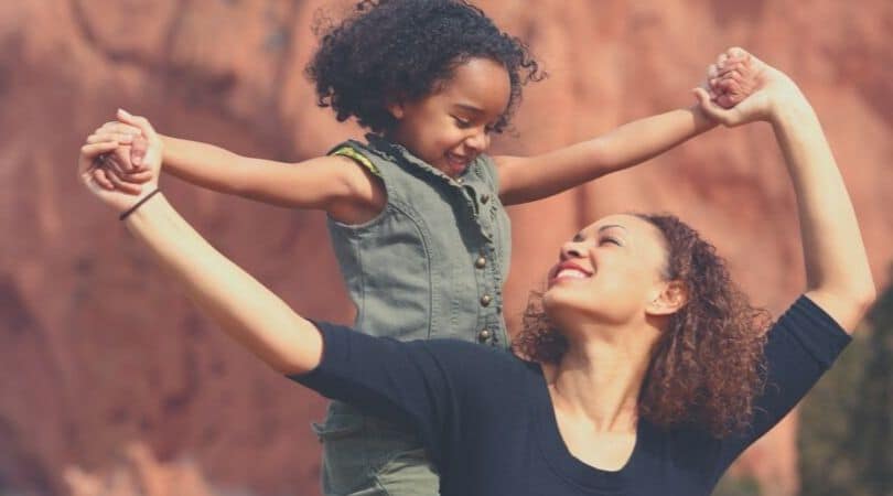 mãe parda, de cabelos ondulados, com um grande sorriso no rosto, com os braços esticados olhando para trás e com os braços abertos apoiando as mãos da filha que está com os braços abertos como um pássaro, olhando sorrindo para baixo em direção aos olhos da mãe, a menina tem aproximadamente cinco anos, negra dos cabelos cacheados e soltos.