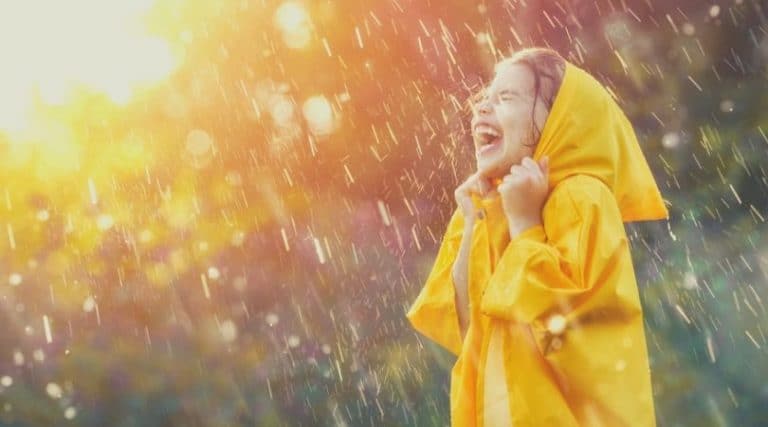 criança de aproximadamente cinco anos, tomando um banho de chuva coberta por sua capa de chuva amarela, no por do sol, bastante alegre com as gotas batendo sobre seu rosto.