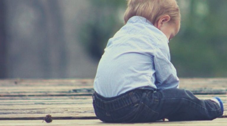 criança de aproximadamente dois anos sentada no chão de madeira, de costas, com a cabeça baixa demonstrando tristeza em sua expressão corporal. t