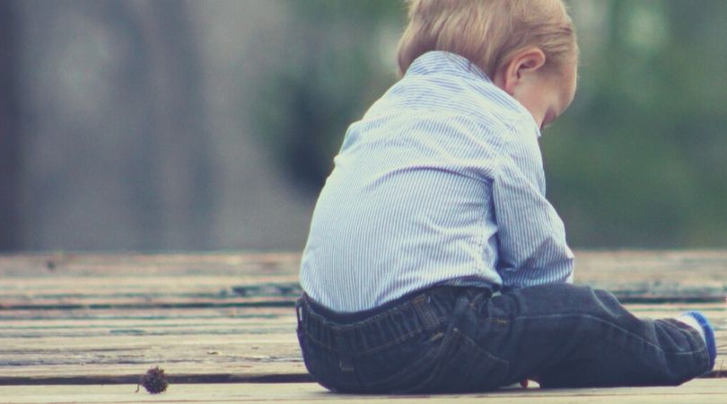 criança de aproximadamente dois anos sentada no chão de madeira, de costas, com a cabeça baixa demonstrando tristeza em sua expressão corporal. t