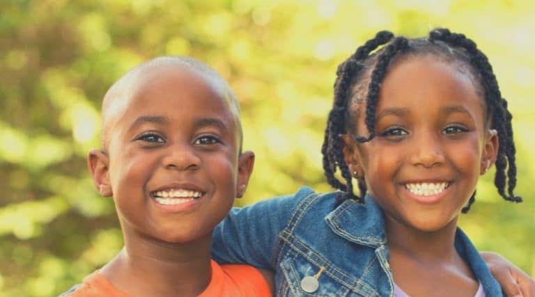 dois irmãos, um menino negro, sorridente de aproximadamente quatro anos e com o cabelo raspado, sendo abraçado pela irmã sorridente, de aproximadamente cinco anor, negra, com o cabelo preso em tranças.